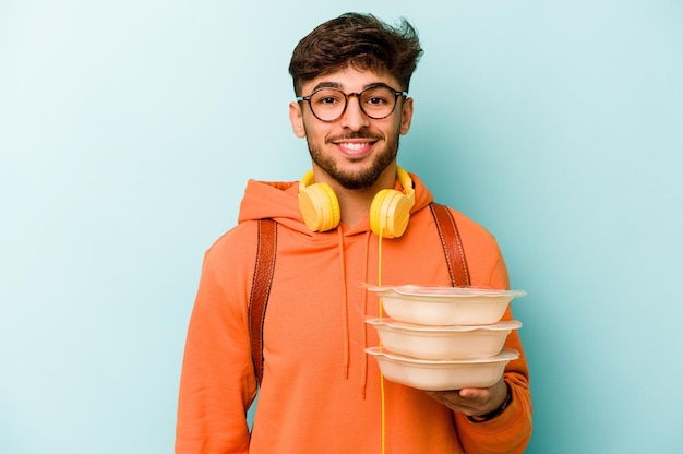 Joven estudiante hispano sosteniendo un tupperware aislado sobre fondo azul feliz sonriendo y alegre