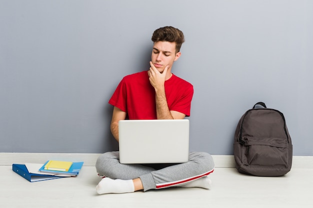 Joven estudiante hombre sentado en el piso de su casa sosteniendo una computadora portátil