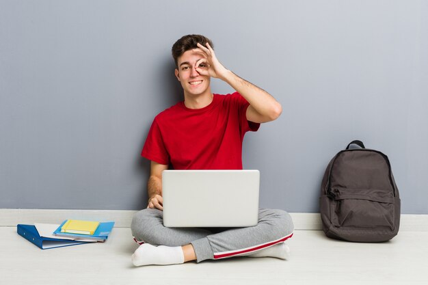 Joven estudiante hombre sentado en el piso de su casa sosteniendo una computadora portátil