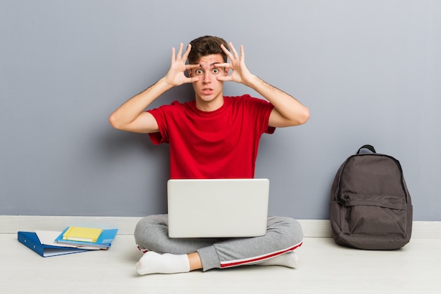 Joven estudiante hombre sentado en el piso de su casa sosteniendo una computadora portátil