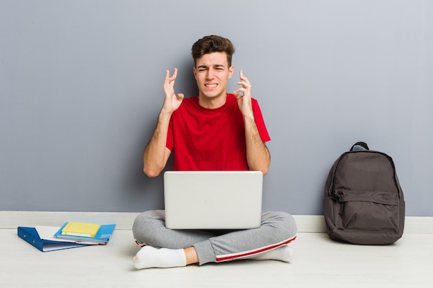 Joven estudiante hombre sentado en el piso de su casa sosteniendo una computadora portátil