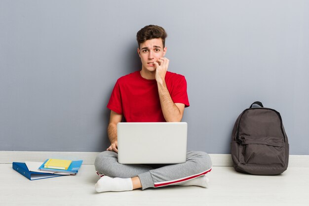 Joven estudiante hombre sentado en el piso de su casa sosteniendo una computadora portátil