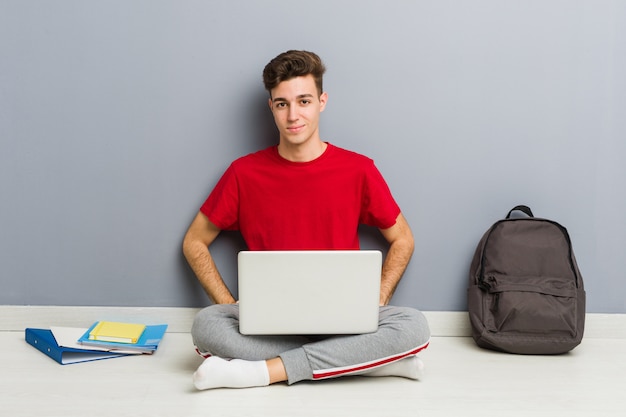 Joven estudiante hombre sentado en el piso de su casa sosteniendo una computadora portátil