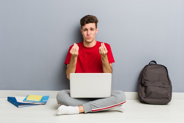 Joven estudiante hombre sentado en el piso de su casa sosteniendo una computadora portátil