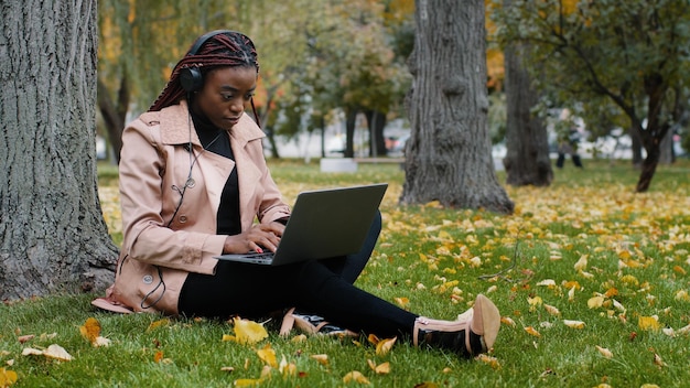 Joven estudiante independiente enfocada sentada en el parque de otoño cerca de un árbol trabajando en una computadora portátil escribiendo