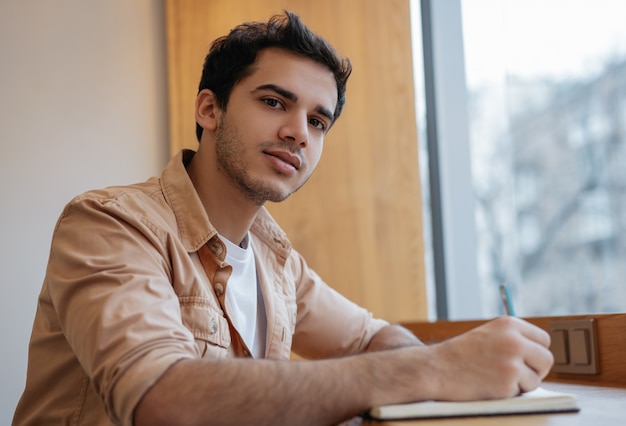 Joven estudiante indio estudiando, preparación de exámenes, tomando notas, mirando a cámara