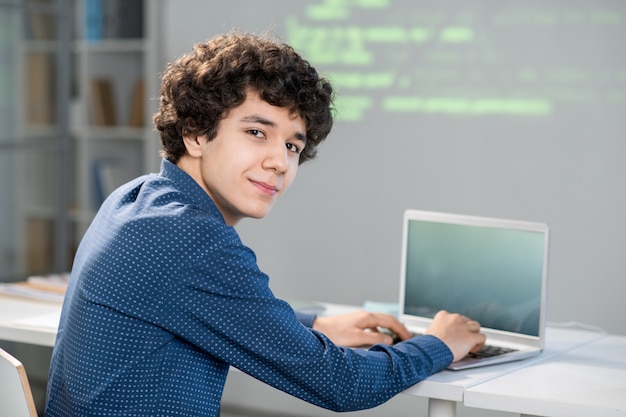 Joven estudiante inteligente con cabello rizado que lo mira mientras está sentado junto al escritorio y trabaja con datos técnicos o prepara una presentación