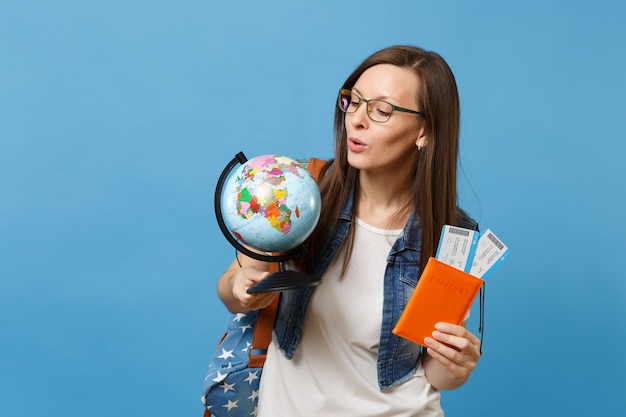 Joven estudiante interesada en gafas mirando guante mundial con pasaporte, billetes de embarque aislado sobre fondo azul. Educación en colegio universitario en el extranjero. Concepto de vuelo de viajes aéreos.