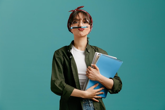 Joven estudiante juguetona con gafas de bandana sosteniendo grandes blocs de notas con ambas manos mirando a la cámara haciendo bigote con bolígrafo aislado en fondo azul