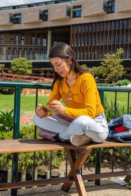 Foto joven estudiante latina con un suéter amarillo y jeans sentada en un banco del campus universitario mientras escribe en un cuaderno