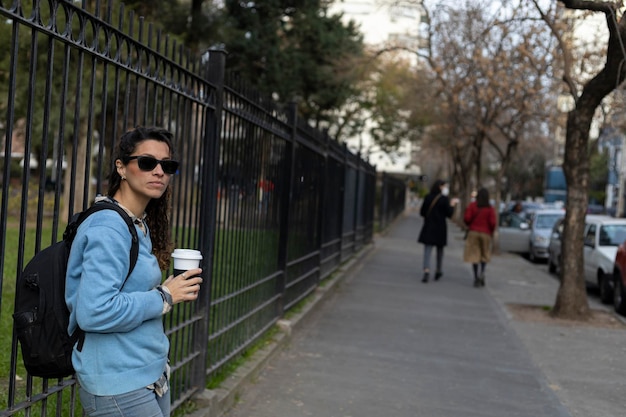 Una joven estudiante latinoamericana espera afuera de la universidad con su vaso de café.