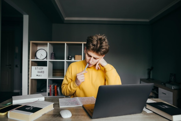 Joven estudiante masculino que estudia en casa con libros y una computadora portátil en el fondo del dormitorio mirando un cuaderno con cara serosa y bolígrafo mordedor Adolescente está estudiando a distancia aprendiendo durante la cuarentena