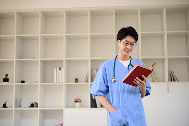 Joven estudiante de medicina asiático inteligente leyendo un libro mientras está de pie en una biblioteca