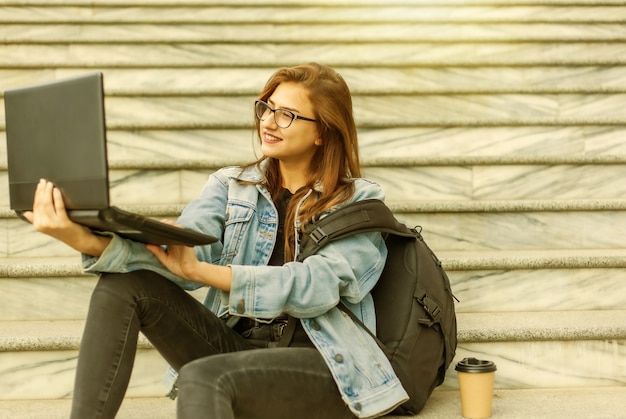 Joven estudiante moderna en chaqueta vaquera y gafas sentado en las escaleras con ordenador portátil. Viendo un video. La educación a distancia. Concepto de juventud moderna.