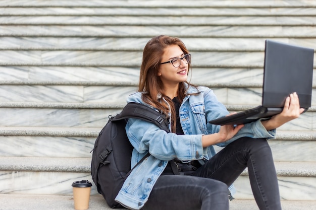 Joven estudiante moderna en chaqueta vaquera y gafas sentado en las escaleras con el portátil. Viendo un video. La educación a distancia. Concepto de juventud moderna.