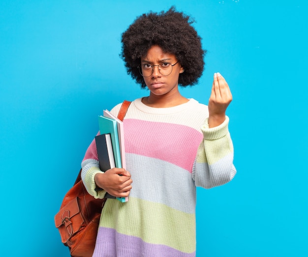 Joven estudiante mujer afro haciendo capice o gesto de dinero, sosteniendo libros