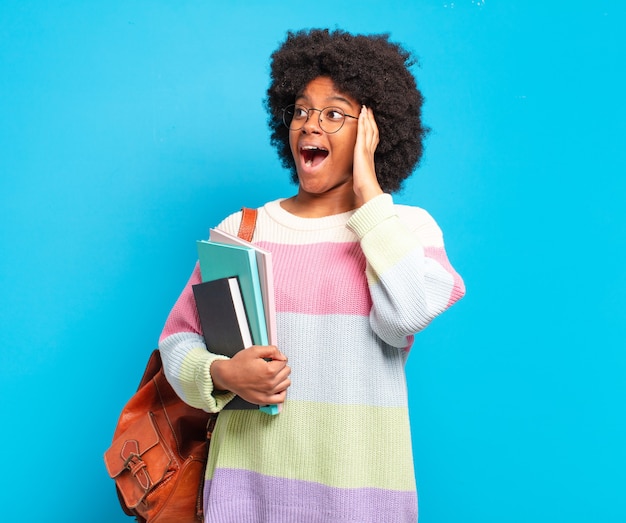 Joven estudiante mujer afro que se siente feliz, emocionada y sorprendida, mirando hacia el lado con ambas manos en la cara