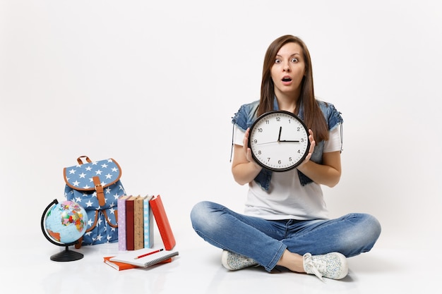 Joven estudiante mujer asustada conmocionada en ropa de mezclilla con reloj despertador sentado cerca del globo, mochila, libros escolares aislados