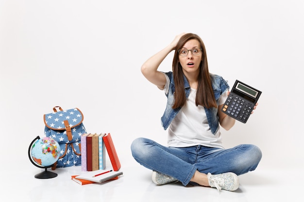 Joven estudiante mujer asustada conmocionada sosteniendo la calculadora aferrándose a la cabeza aprendiendo matemáticas sentado cerca del globo, mochila, libros escolares aislados