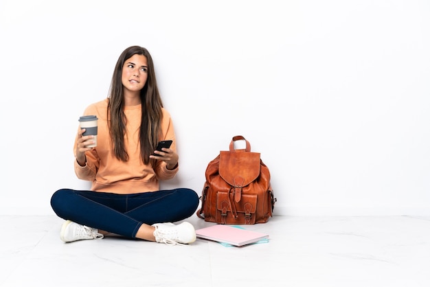 Joven estudiante mujer brasileña sentada en el suelo sosteniendo un café para llevar y un móvil mientras piensa en algo