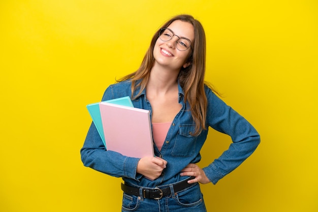 Joven estudiante mujer caucásica aislada de fondo amarillo posando con los brazos en la cadera y sonriendo