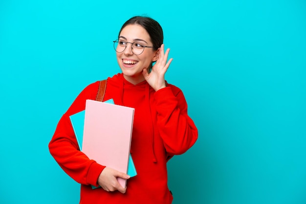 Joven estudiante mujer caucásica aislada de fondo azul escuchando algo poniendo la mano en la oreja