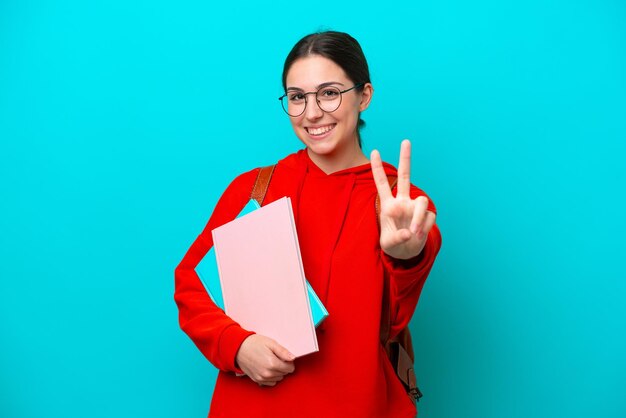 Joven estudiante mujer caucásica aislada de fondo azul sonriendo y mostrando el signo de la victoria