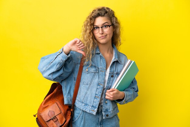 Joven estudiante mujer caucásica aislada sobre fondo amarillo mostrando el pulgar hacia abajo con expresión negativa