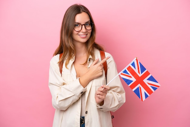 Joven estudiante mujer caucásica sosteniendo una bandera del Reino Unido aislada en un fondo rosa apuntando hacia un lado para presentar un producto