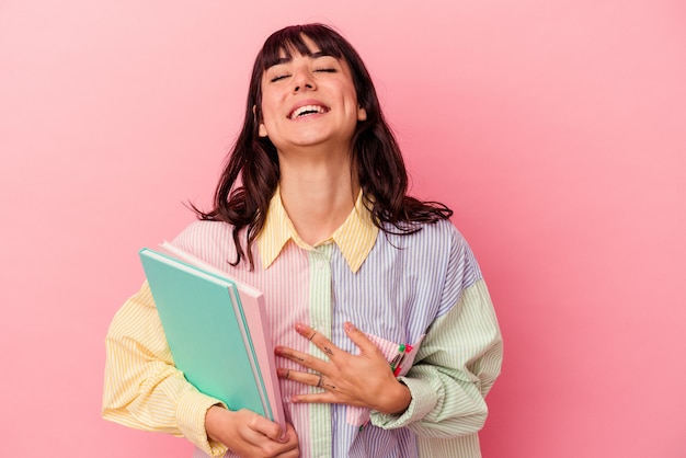 Joven estudiante mujer caucásica sosteniendo libros aislados sobre fondo rosa se ríe a carcajadas manteniendo la mano en el pecho.