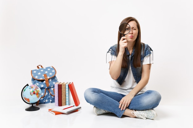Joven estudiante mujer interesada desconcertado sosteniendo mirando lupa sentado cerca del globo, mochila, libros escolares aislados