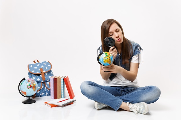 Joven estudiante mujer interesada inteligente mirando globo con lupa aprendiendo sentado cerca de la mochila, libros escolares aislados