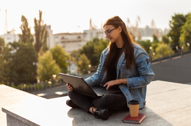 Joven estudiante mujer moderna en una chaqueta de mezclilla sentado y utiliza el portátil al aire libre. La educación a distancia. Concepto de juventud moderna.