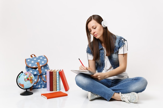 Joven estudiante mujer pensativa en auriculares escuchando música escribiendo notas en el cuaderno sentado cerca de la mochila del globo, libros escolares aislados