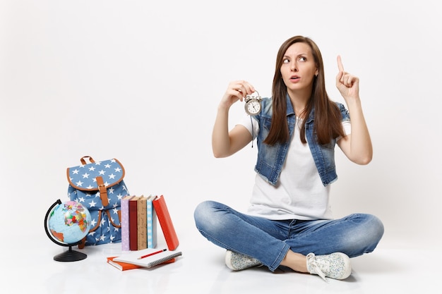 Joven estudiante mujer preocupada apuntando con el dedo índice hacia arriba sosteniendo el reloj despertador sentado cerca del globo, mochila, libros escolares aislados en la pared blanca