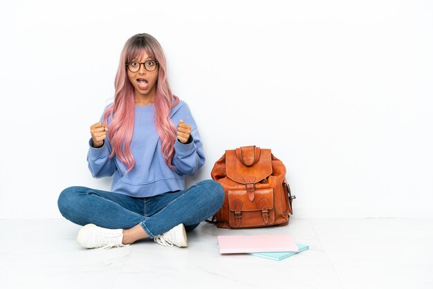 Joven estudiante mujer de raza mixta con cabello rosado sentado en el piso aislado sobre fondo blanco celebrando una victoria en la posición ganadora