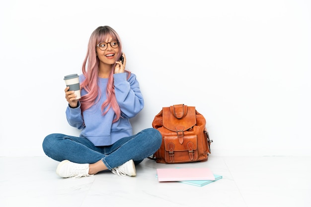 Joven estudiante mujer de raza mixta con cabello rosado sentado en el piso aislado sobre fondo blanco sosteniendo café para llevar y un móvil