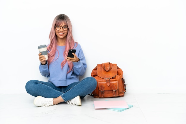 Joven estudiante mujer de raza mixta con cabello rosado sentado en el piso aislado sobre fondo blanco sosteniendo café para llevar y un móvil