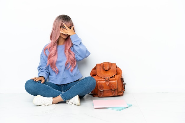 Joven estudiante mujer de raza mixta con cabello rosado sentado en el suelo aislado sobre fondo blanco cubriendo los ojos con las manos y sonriendo