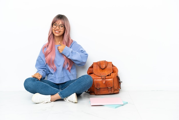 Foto joven estudiante mujer de raza mixta con cabello rosado sentado en el suelo aislado sobre fondo blanco dando un pulgar hacia arriba gesto