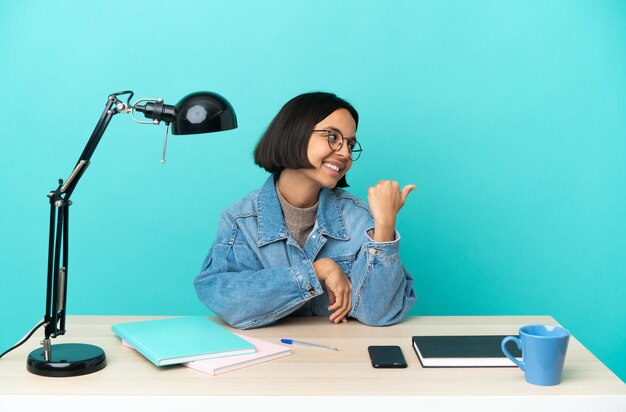 Joven estudiante mujer de raza mixta estudiando sobre una mesa apuntando hacia el lado para presentar un producto