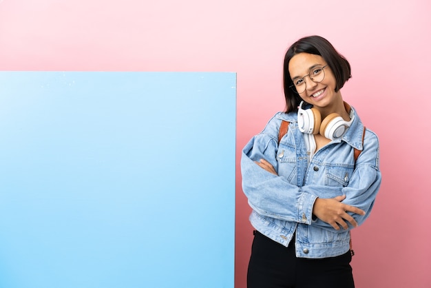 Joven estudiante mujer de raza mixta con una gran pancarta sobre una pared aislada con los brazos cruzados y mirando hacia adelante