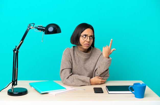 Joven estudiante mujer de raza mixta que estudia en una mesa haciendo el gesto de locura poniendo el dedo en la cabeza