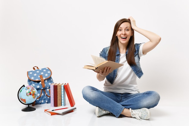 Joven estudiante mujer riendo sorprendido en ropa de mezclilla mantenga la mano de mantenimiento de libros cerca de la cabeza sentarse cerca del globo, mochila, libros escolares aislados