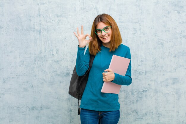 Joven estudiante mujer sintiéndose feliz, relajada y satisfecha, mostrando aprobación con gesto bien, sonriendo contra la pared del grunge