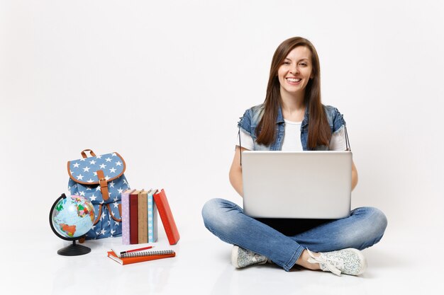 Joven estudiante mujer sonriente alegre casual sosteniendo usando una computadora portátil sentado cerca del globo, mochila, libros escolares aislados en la pared blanca