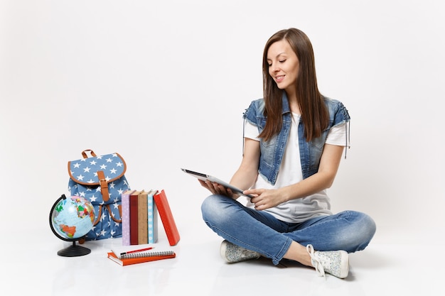 Joven estudiante mujer sonriente casual en ropa de mezclilla con tablet pc sentado cerca de libros de escuela de mochila de globo aislado