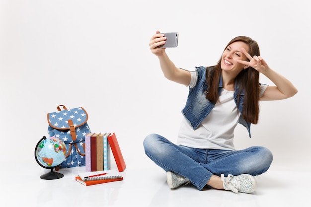 Foto joven estudiante mujer sonriente haciendo tomar selfie foto en teléfono móvil mostrando el signo de la victoria cerca del globo, mochila, libros escolares aislados en la pared blanca