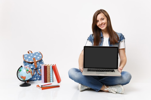 Joven estudiante mujer sonriente sosteniendo una computadora portátil con pantalla vacía en blanco negro sentado cerca de los libros escolares de la mochila del globo aislado