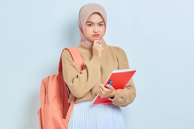 Una joven estudiante musulmana asiática pensativa con suéter marrón con mochila sosteniendo un libro y pensando en algo aislado en el concepto de regreso a la escuela de fondo blanco